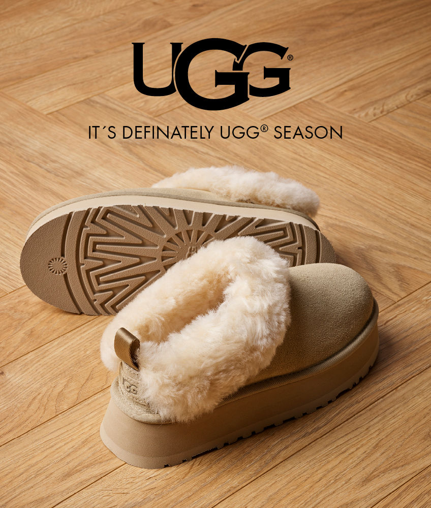 Beige UGG-tøfler med myk, hvit pelskant på tregulv. UGG-logo og teksten "IT'S DEFINATELY UGG SEASON" vises øverst.
