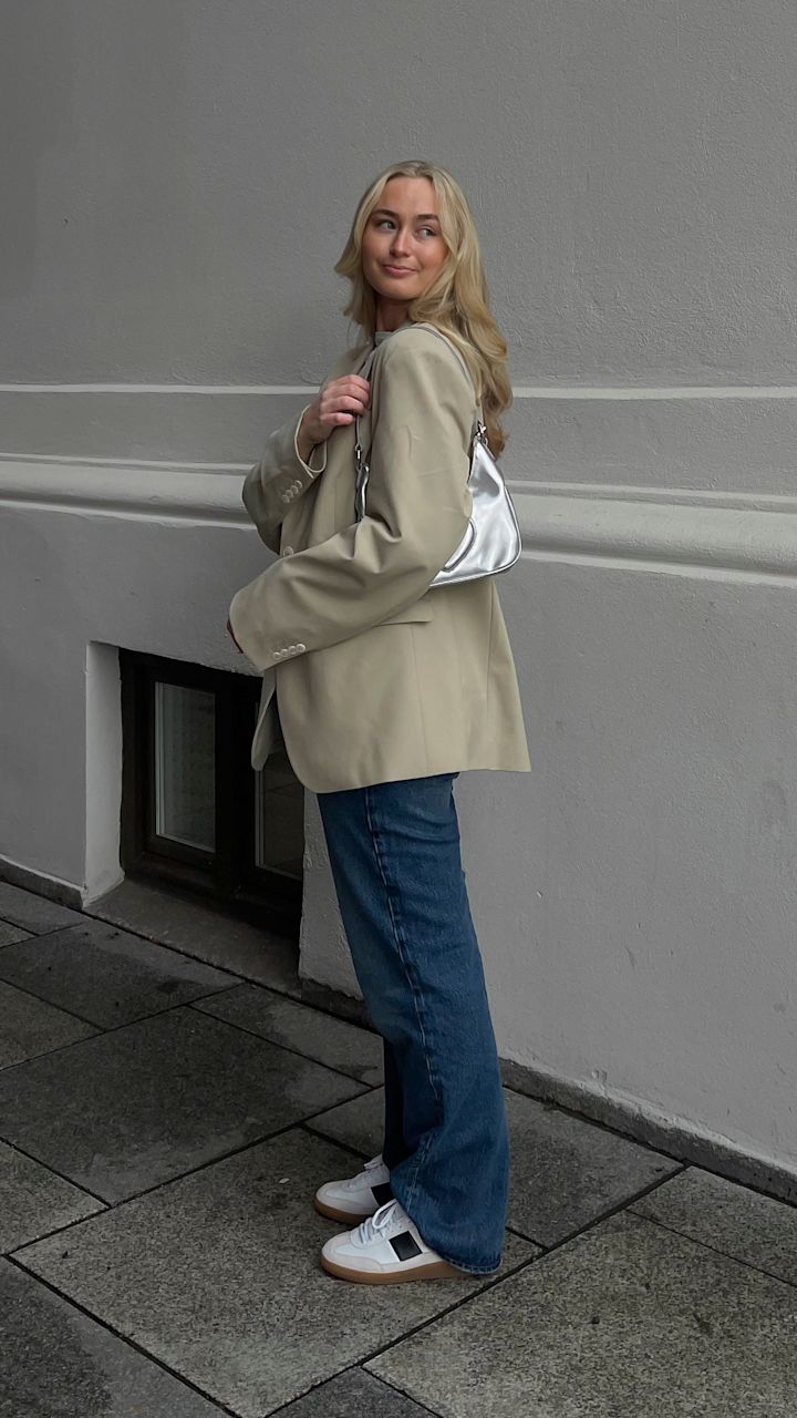 Ung kvinne med blondt hår som står utendørs ved en grå vegg, iført beige blazer, blå jeans, hvite sneakers og bærer en sølvfarget veske over skulderen.