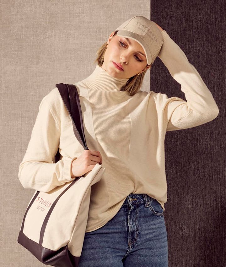 Person med beige caps og kremfarget genser holder en stoffveske med brune detaljer