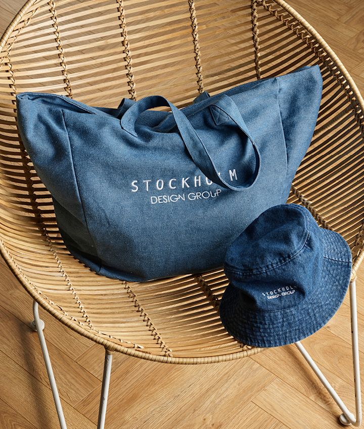 Blå denim tote bag og bucket hat med "Stockholm Design Group"-logo på en rottingstol med tregulv.