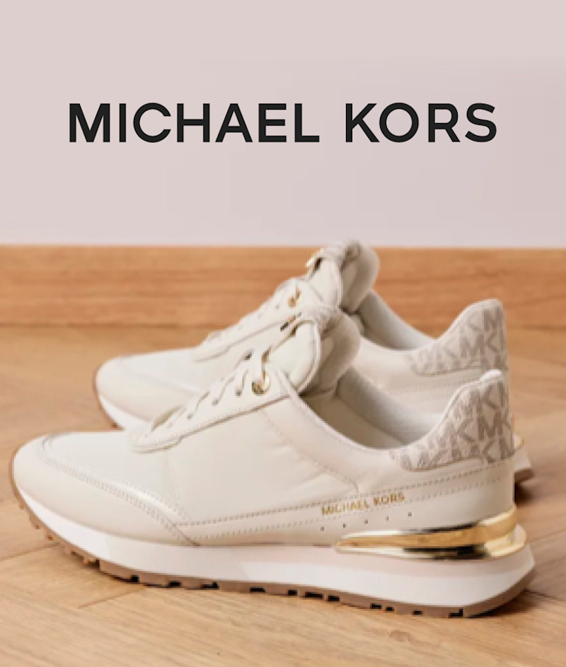 Hvite Michael Kors sneakers med gulldetaljer og merkelogo på hælen, plassert på tregulv med merkenavnet øverst i bildet.