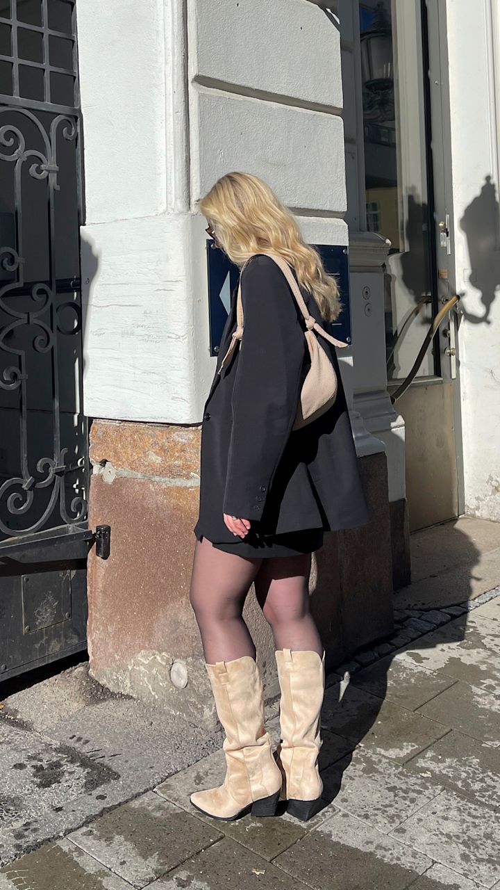 Kvinne med blondt hår går ved hjørnet av en bygning iført svart blazer, kort skjørt, svarte strømpebukser og beige støvler, med en beige veske over skulderen.