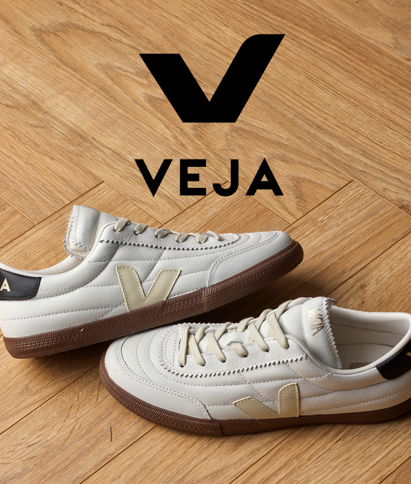Hvite VEJA sneakers med beige V-logo og brun gummisåle på tregulv, med merkets logo synlig øverst.