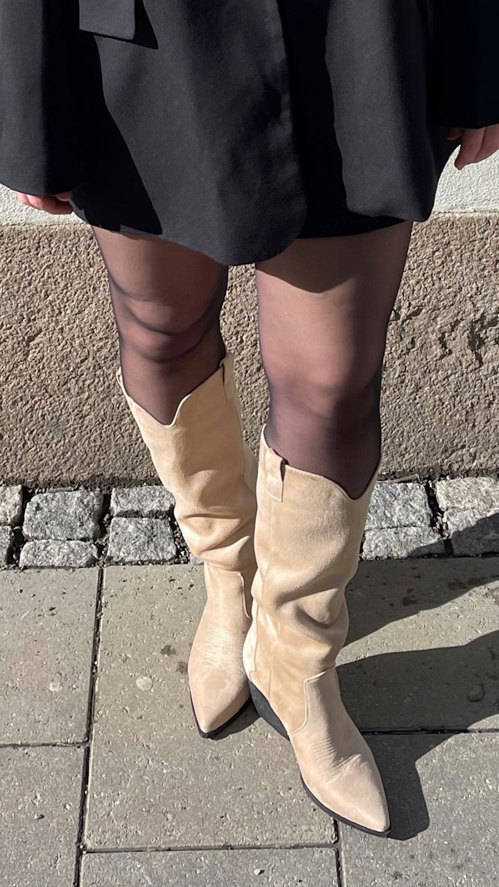 Bein iført svarte strømpebukser og beige skinnstøvler med spiss tupp, stående på brosteins- og betongfortau ved en murvegg.