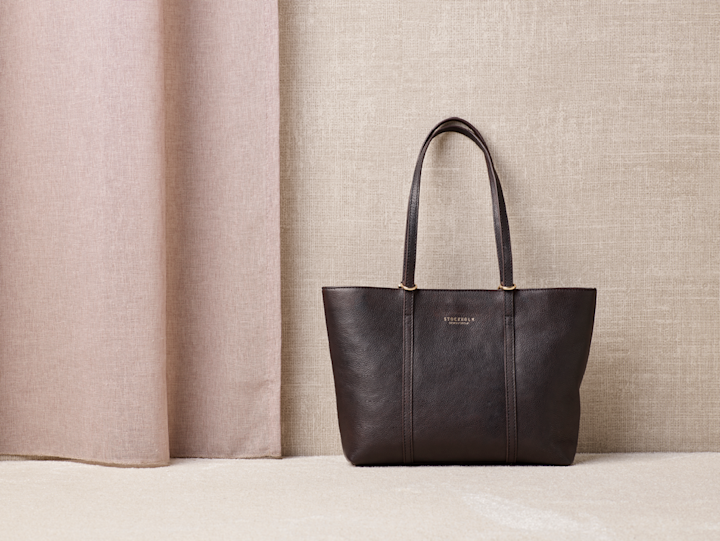 Elegant brun skinnveske med lange håndtak mot en nøytral beige bakgrunn med tekstilgardiner.