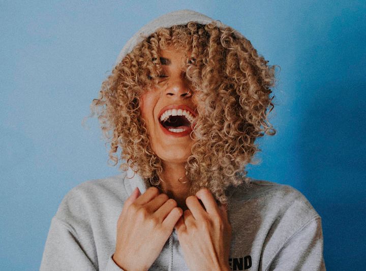 Ung kvinne med krøllete hår og grå hettegenser ler høyt foran blå bakgrunn.