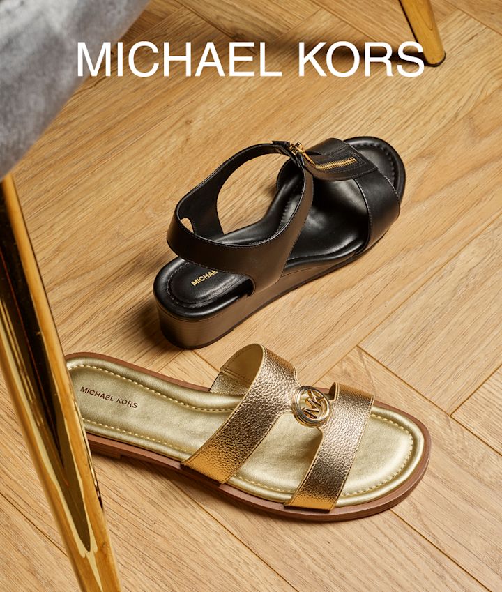 To Michael Kors-sandaler på tregulv: en sort kilehæl-sandal og en flat gull slippers med MK-logo.