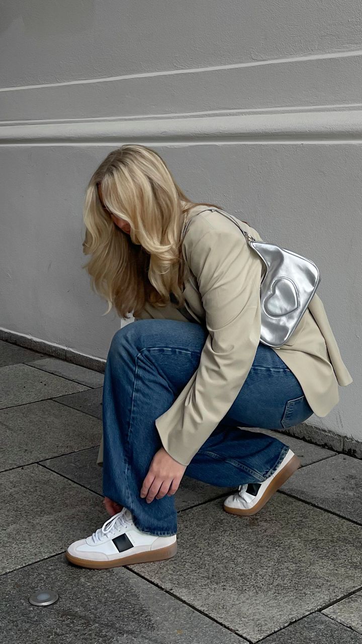 Kvinne med blondt hår bøyer seg ned for å knyte hvite sneakers med svart detalj på grå steinlagt gate, iført beige blazer, blå jeans og sølvfarget veske med hjerte.