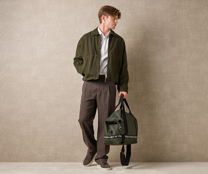 Person i olivengrønn jakke og brune bukser med matchende brune sneakers, holder en mørkegrønn veske mot beige bakgrunn.