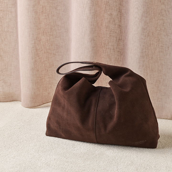 Elegant mørkebrun semsket skinnveske med håndtak, plassert på et kremfarget teppe mot lyse gardiner.