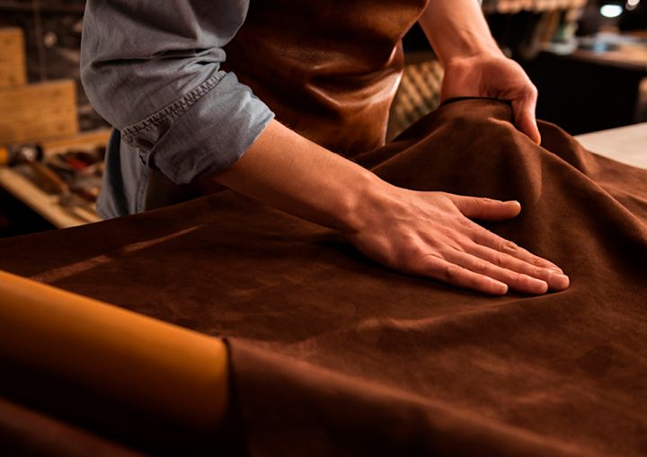 Hender som berører og glatter ut et stykke brunt lær på et arbeidsbord i et verksted.