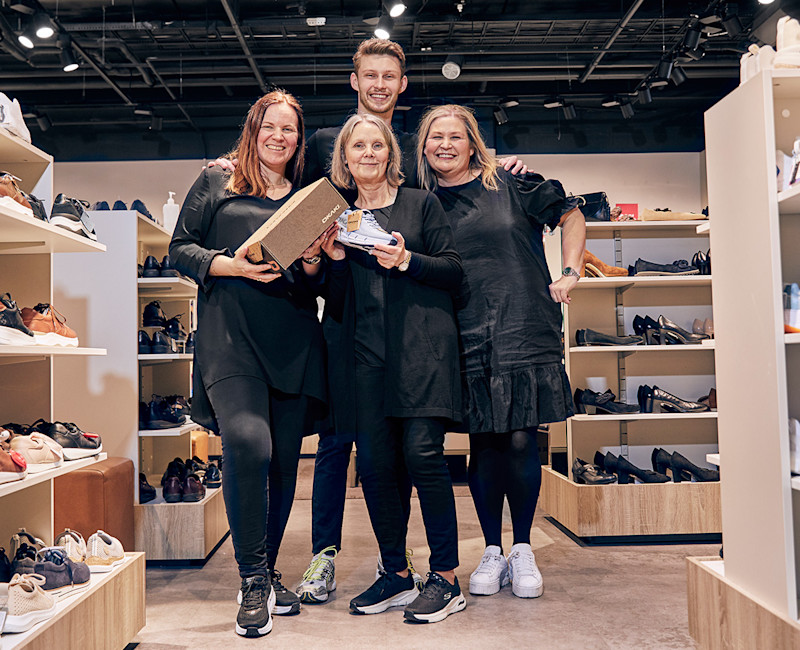 Fire smilende personer står sammen i en skobutikk, to av dem holder en skoeske og en sko, omgitt av hyller med ulike sko.