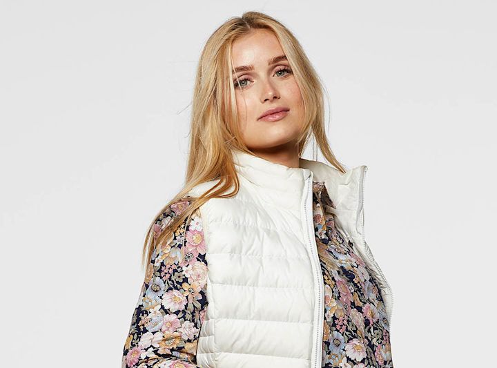 Ung kvinne med langt blondt hår som har på seg en hvit vattert vest over en blomstermønstret skjorte, stående mot en lys bakgrunn.