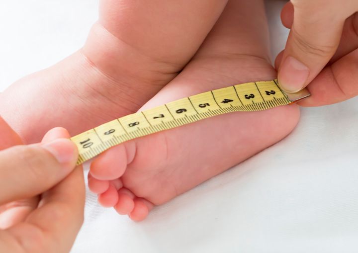 Babyfot blir målt med et målebånd holdt av to hender mot en hvit bakgrunn.