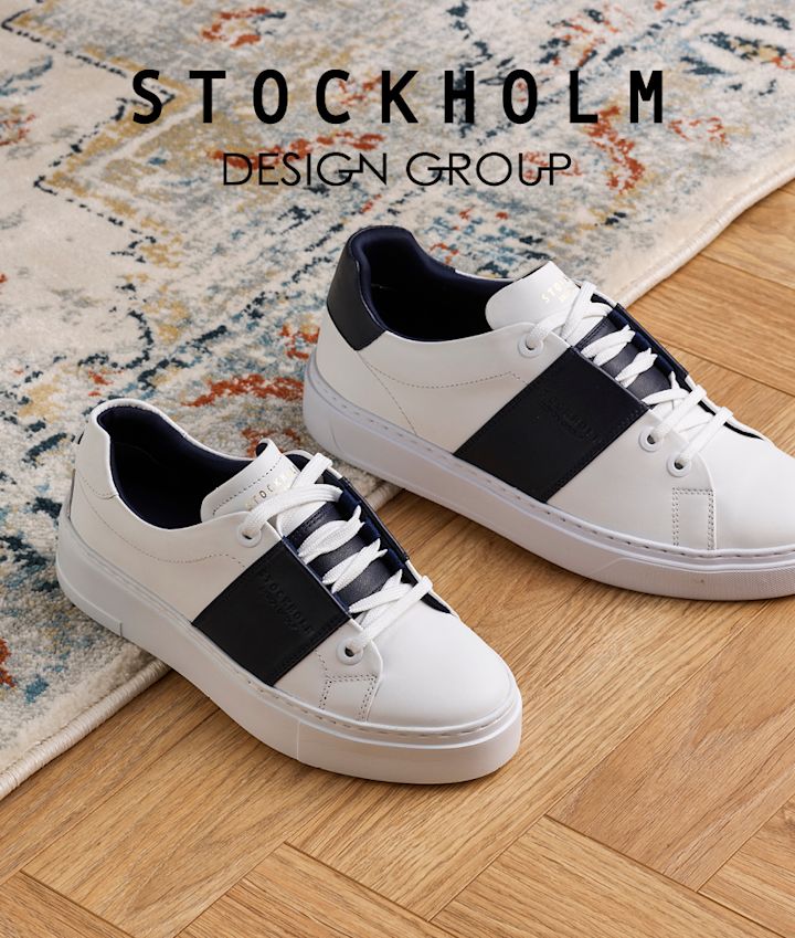 Hvite sneakers med blått sidefelt fra Stockholm Design Group på tregulv ved siden av et mønstret teppe.