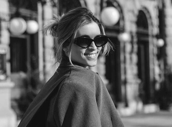 Kvinne med solbriller smiler og ser over skulderen i urbant miljø med bygninger i bakgrunnen.