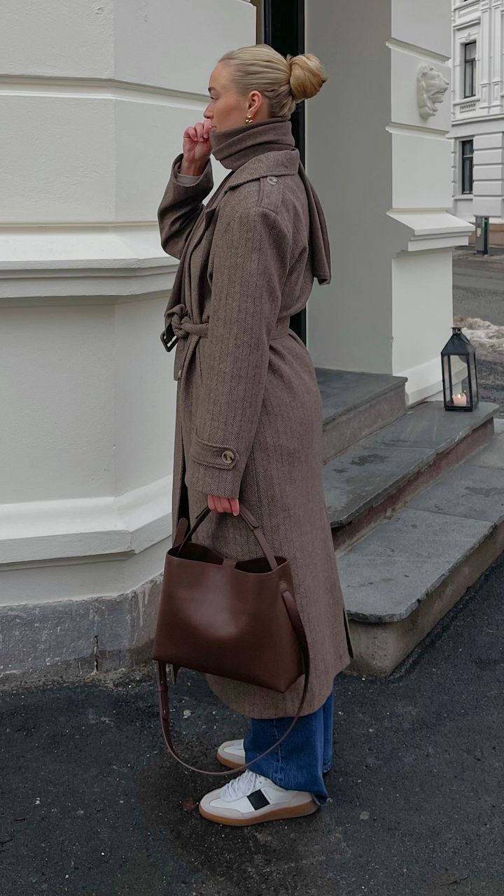 Kvinne med blond hår i knute iført lang brun kåpe, blå jeans og hvite sneakers, holder brun skinnveske og står ved trapp utenfor hvit bygning.