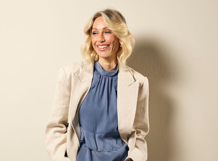 Kvinne med blondt bølget hår smiler og ser til høyre, iført blå bluse og beige blazer, foran en lys vegg.