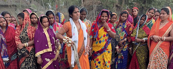 En gruppe kvinner i fargerike tradisjonelle sarier står sammen utendørs, noen smiler mens andre ser alvorlige ut, omgitt av trær og naturlig bakgrunn.