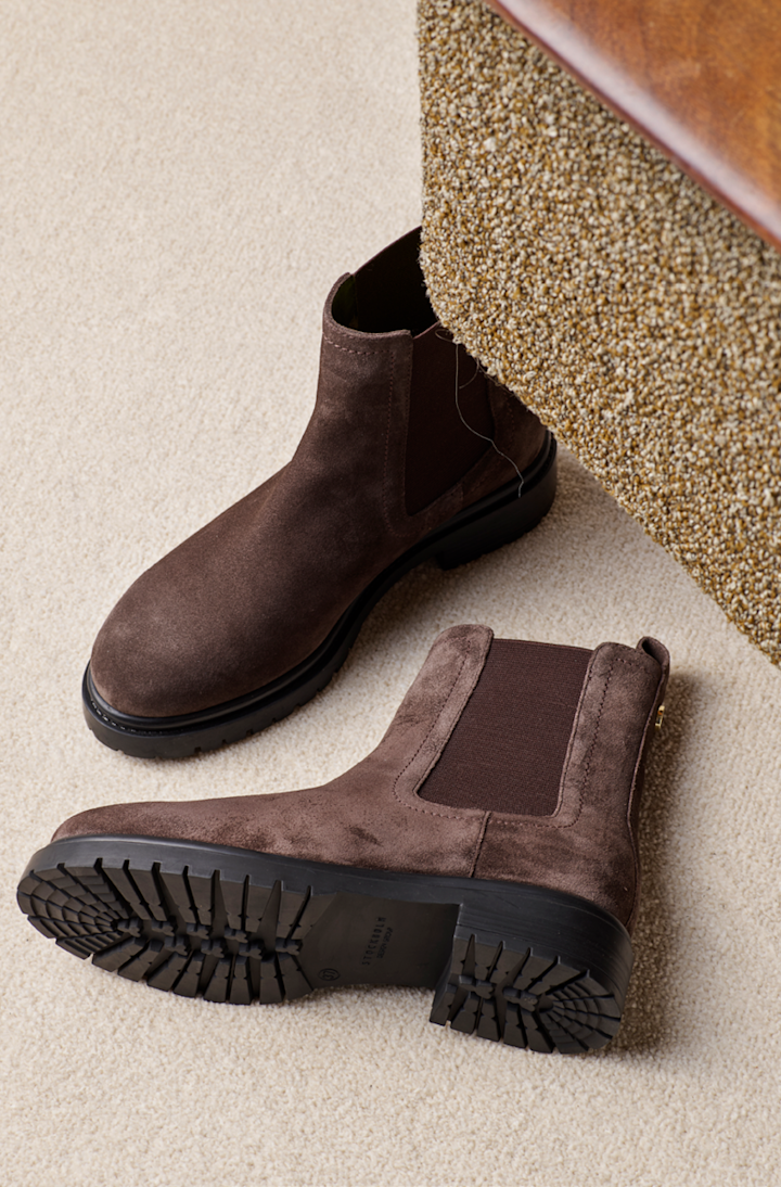 Brune semsket chelsea-støvler med elastiske sider og kraftig sort såle, plassert på beige teppe under en stol.